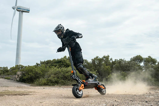 Rider in full protective gear drifting on Freeboy H9 black‑orange off‑road electric scooter on gravel terrain with wind turbine and green shrubs in background